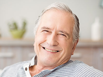 The image shows a smiling older man with gray hair, wearing a blue shirt, sitting comfortably in a home setting with his arm resting on a table.